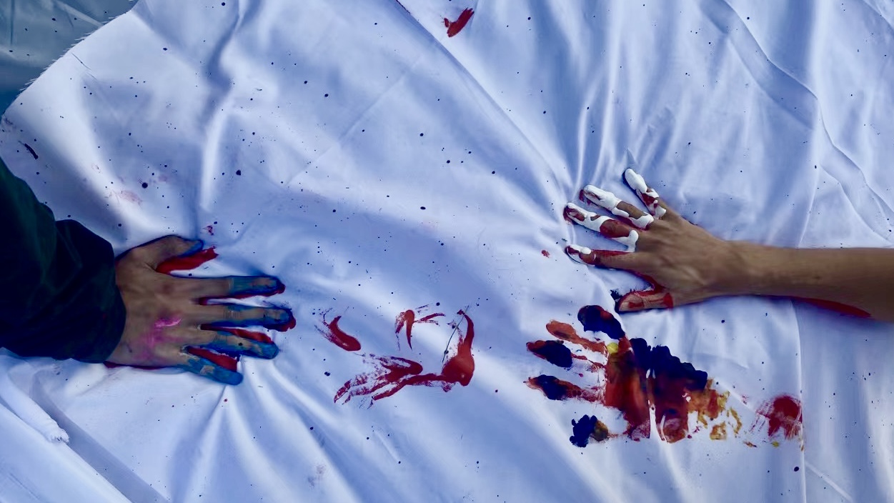 Close-up of two painted hands pressing onto a white mat, leaving red, blue, and purple marks during an outdoor color workshop.