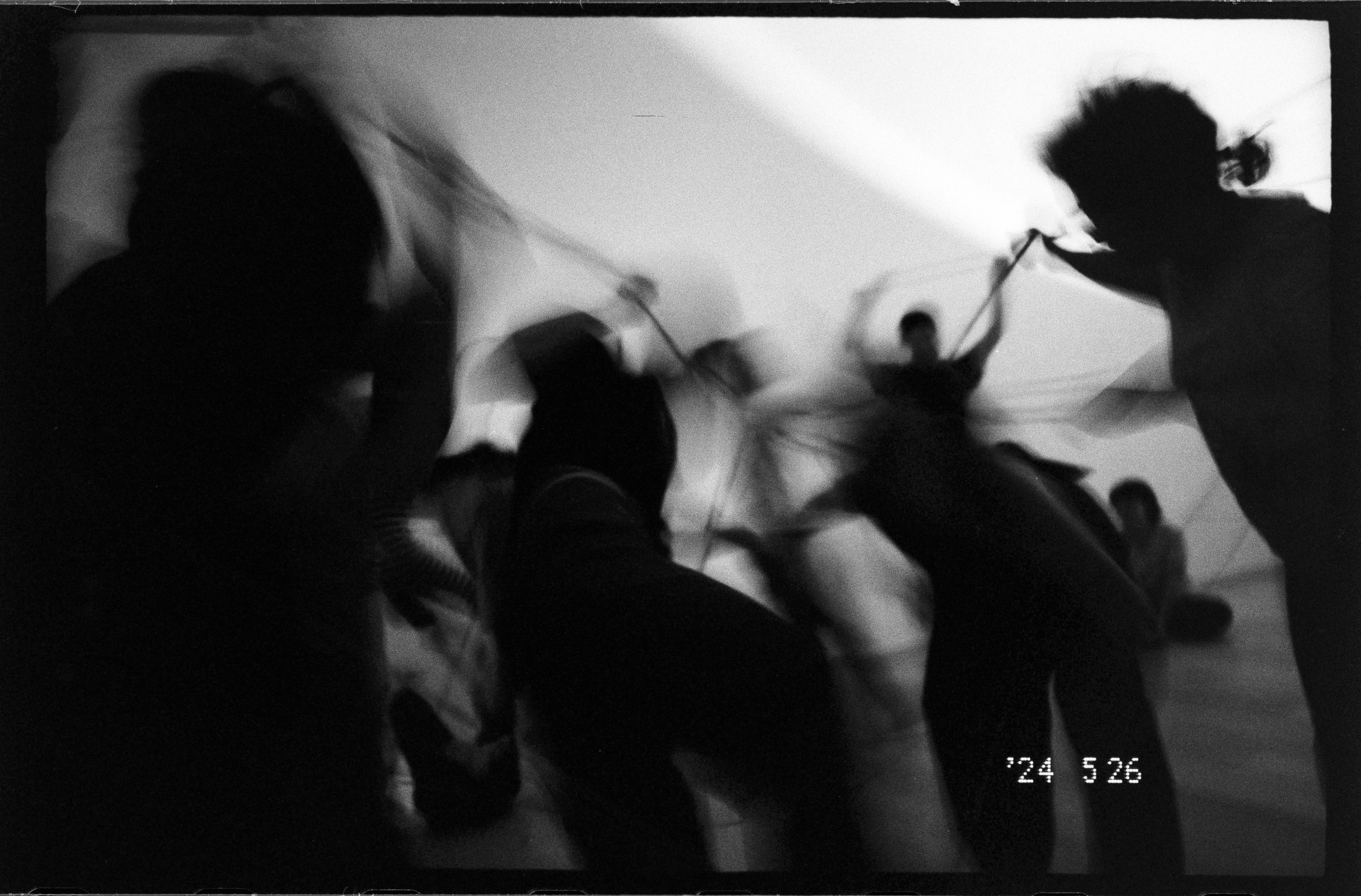 A black-and-white workshop image showing several blurred figures moving with long lines in a dim room, creating a sense of motion and connection.